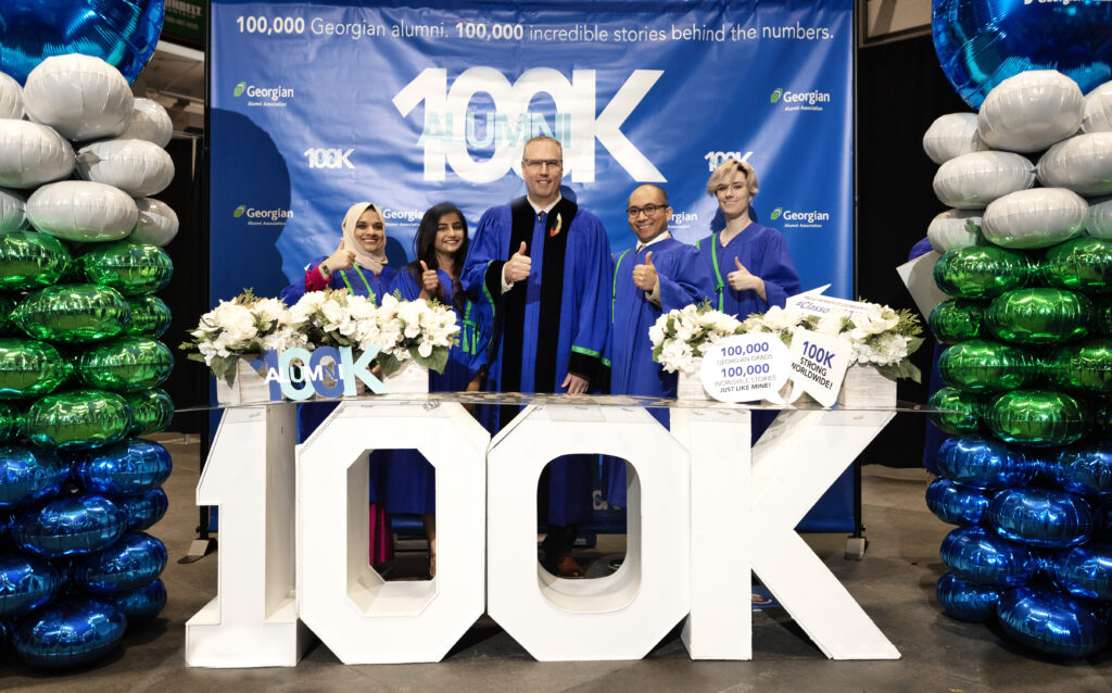 Five people wearing convocation robes stand together in front of a large 100K marquee sign.