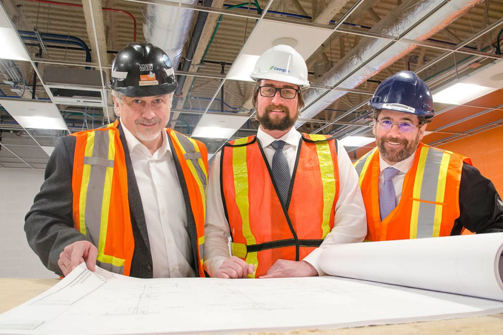 Three men in hard hats and reflective vests standing in front of blueprints