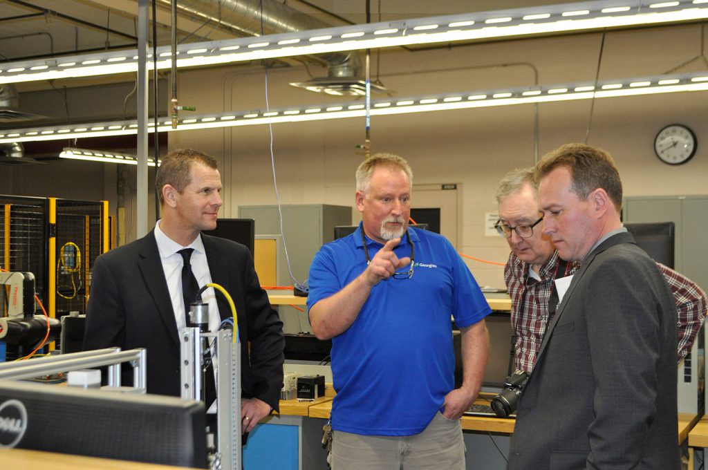 a man in a blue shirt talking to three men in suits in an engineering lab