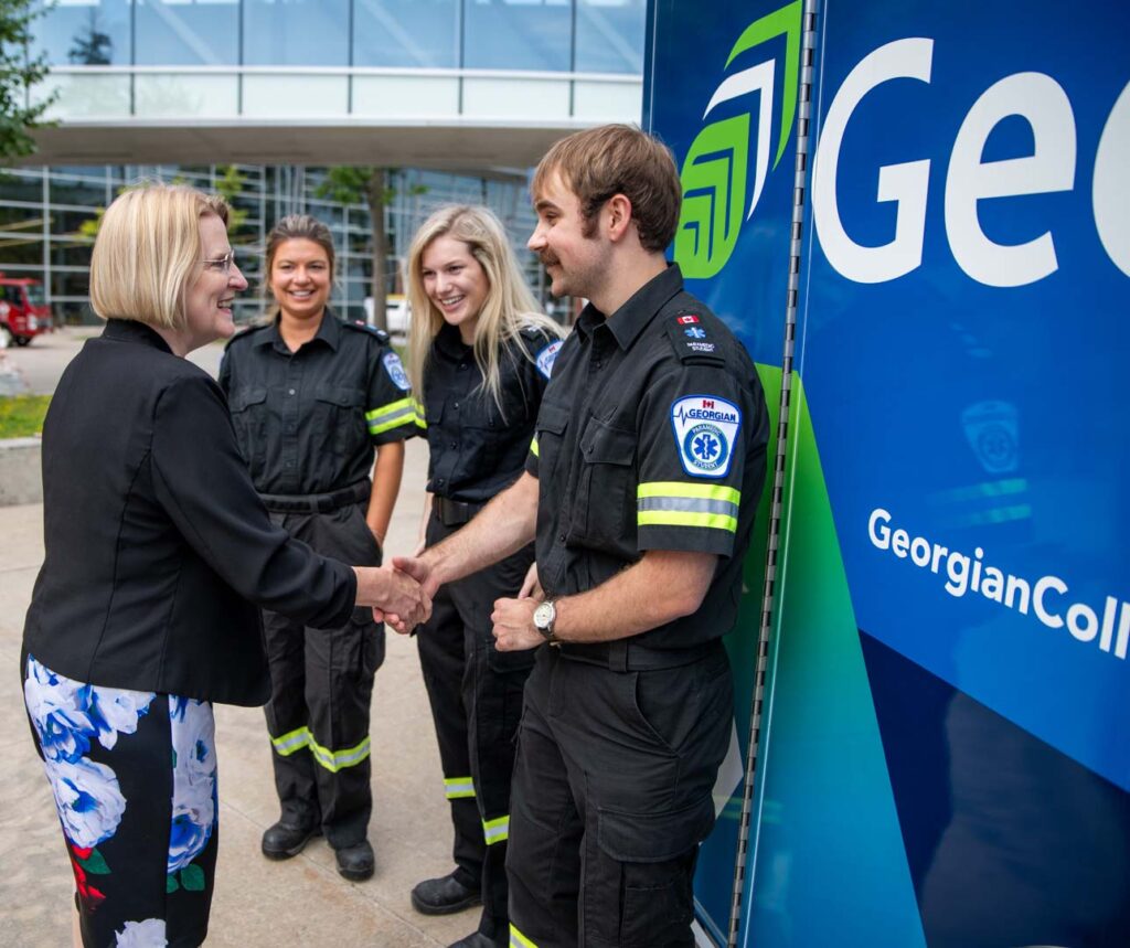 Minister Sylvia Jones shakes the hand of a paramedic student standing in front of an ambulance training vehicle, while two students stand in the background.