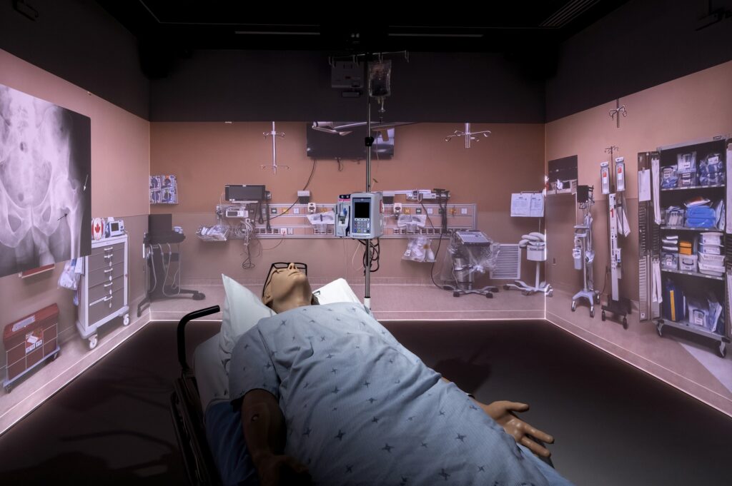 A high fidelity simulator lays in a hospital bed inside a simulation room with the background of an operating room projected on the walls.