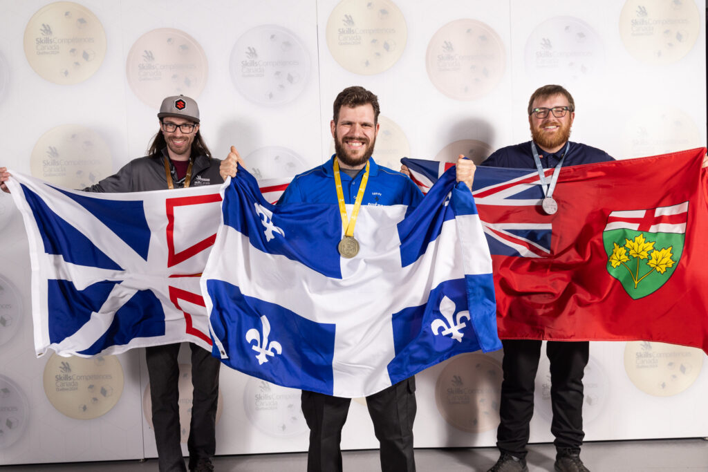 Three people stand with Canadian province flags with medals around their necks.
