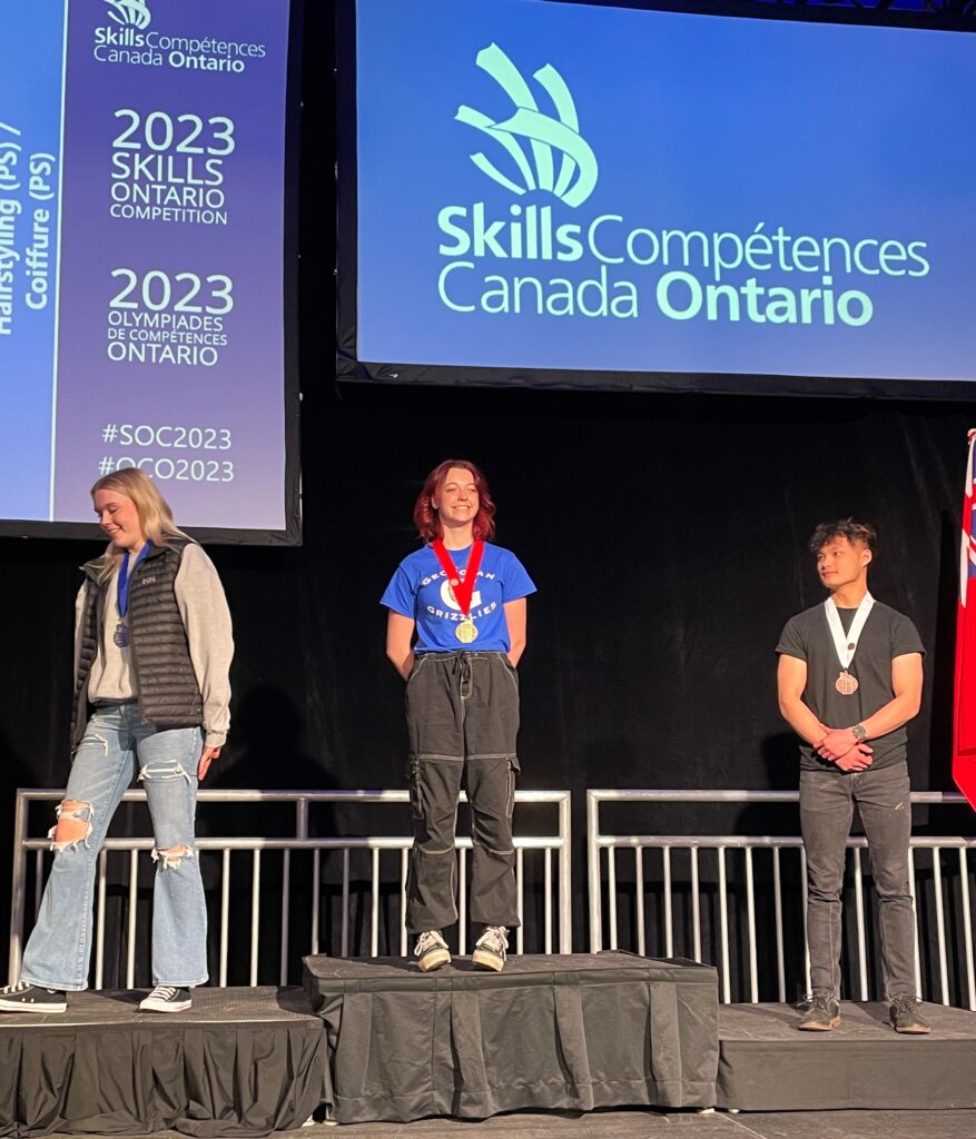 Three people stand on a black podium with gold, silver and bronze medals with the Skills Ontario sign behind them.