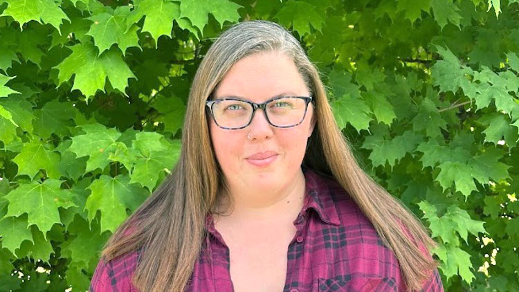Amber Cooper, a Business Administration Human Resources summer student is dressed in a maroon plaid shirt and is standing in front of green trees