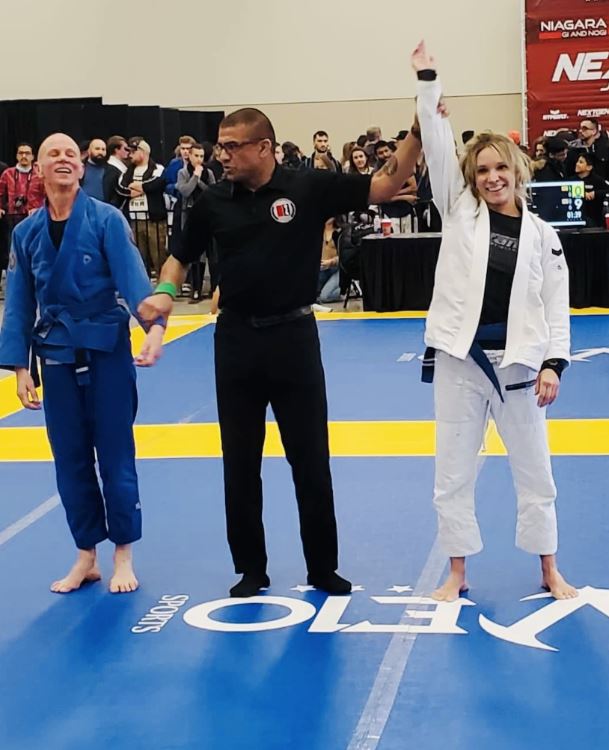 Three adults wearing martial arts uniforms stand on a mat, with the person in the middle raising up an arm of one of them.