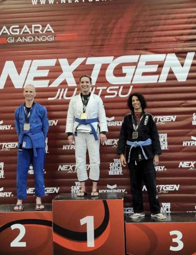 Three people dressed in jiu-jitsu uniforms stand on a winner's podium, with the first-place person on the tallest section. Text: NextGen Jiu Jitsu 