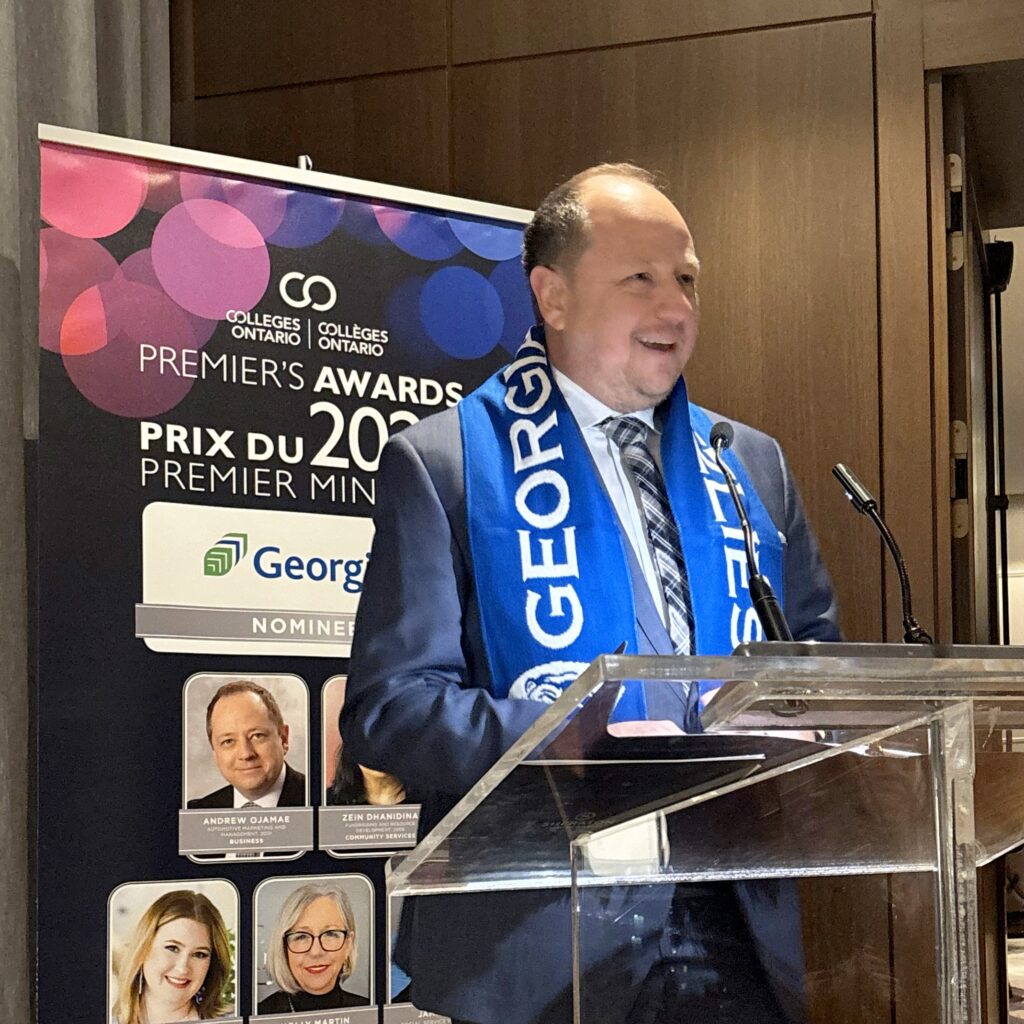 Georgian College alumnus Andrew Ojamae speaks at a podium at the 2024 Premier's Awards, held in Toronto. He's wearing a blue Georgian Grizzlies scarf around his neck and is wearing a dark neutral suit.