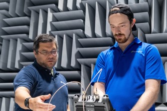 Two researchers examining electronic testing equipment inside an anechoic chamber lined with sound-absorbing panels.