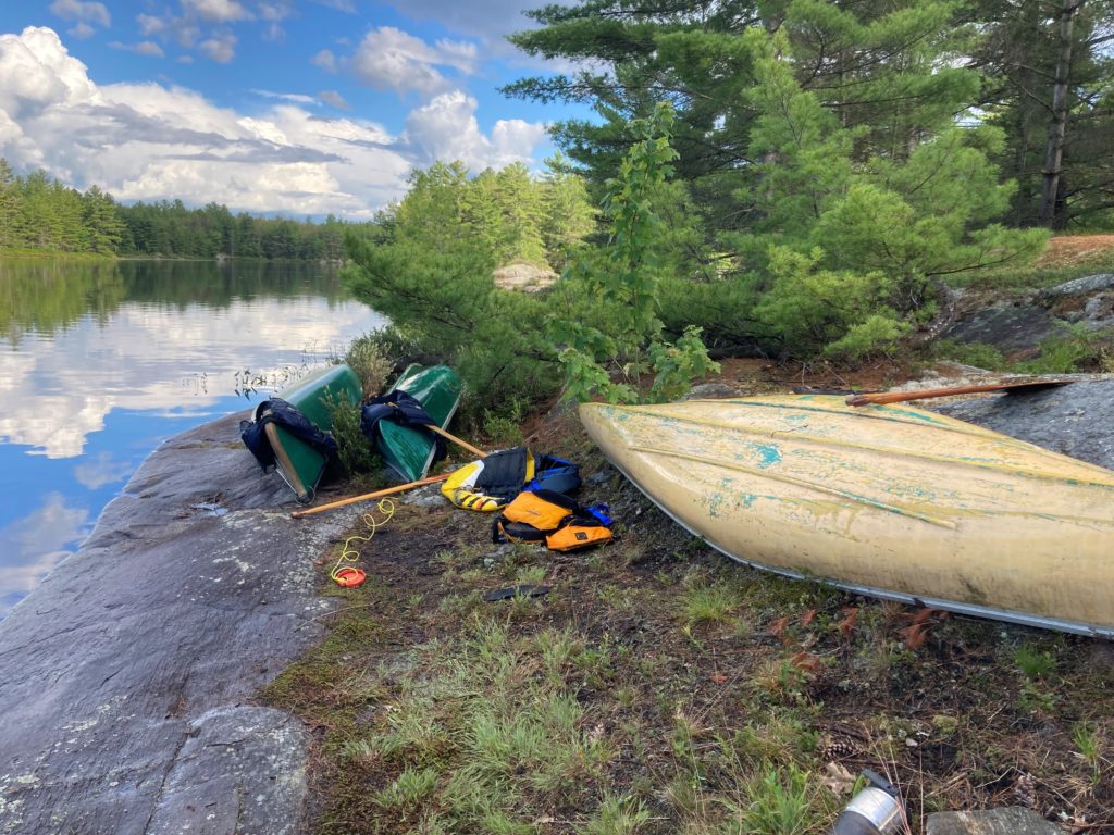 Canoes and lifejackets on a grassy bank next to a lake in the woods.
