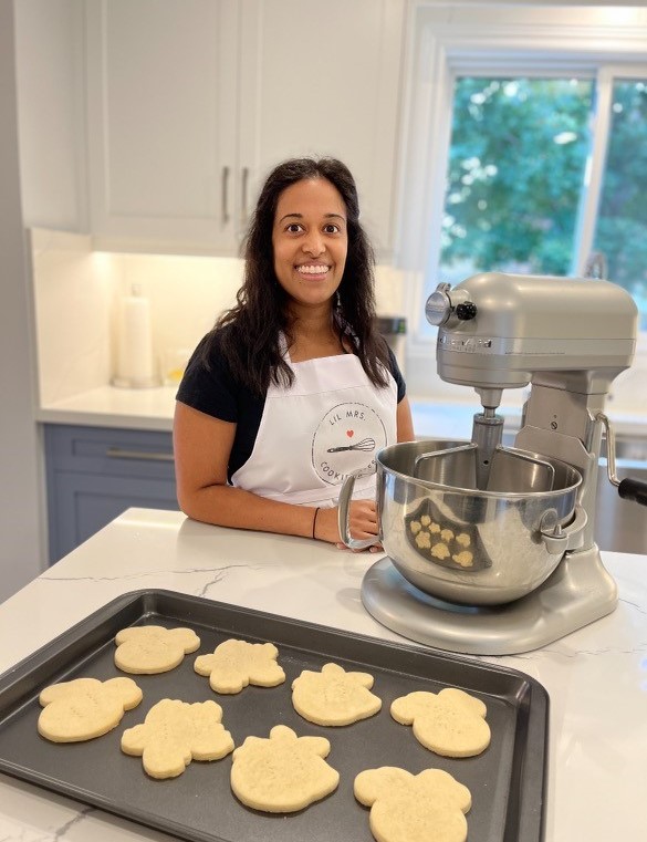 A person with long, brown hair, white apron and black T-shirt, smiles in a kitchen next to a kitchen mixer and a tray of sugar cookies.