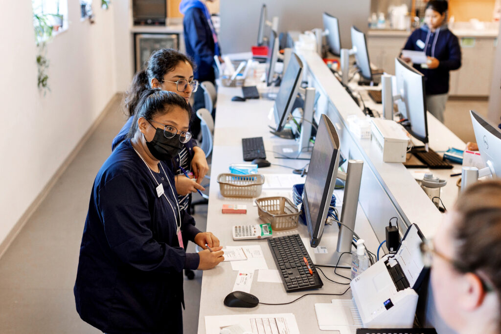 A woman wearing a black mask and a dark blue lab coat writes notes as she looks at another person. Computers surround them in a lab space.
