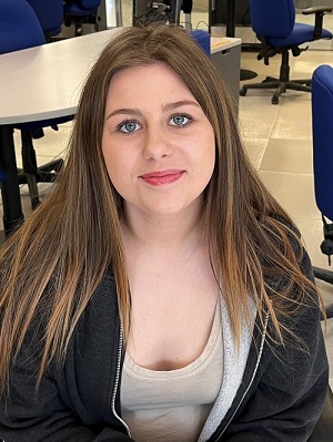Bailey Simone - grad of School Within a College, young woman sitting down with long brown hair, warm smile, in a classroom wearing zip up hoodie and t-shirt