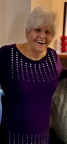 A person with short, white hair and wearing a purple sweater, smiles.