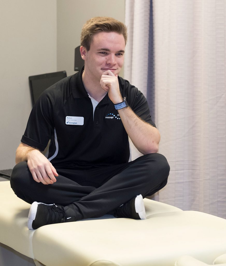 Young male sitting cross-legged on a massage table wearing black scrubs