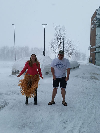 A girl wearing a grass skirt and a guy in shorts and t-shirt stand on a snowy sidewalk.