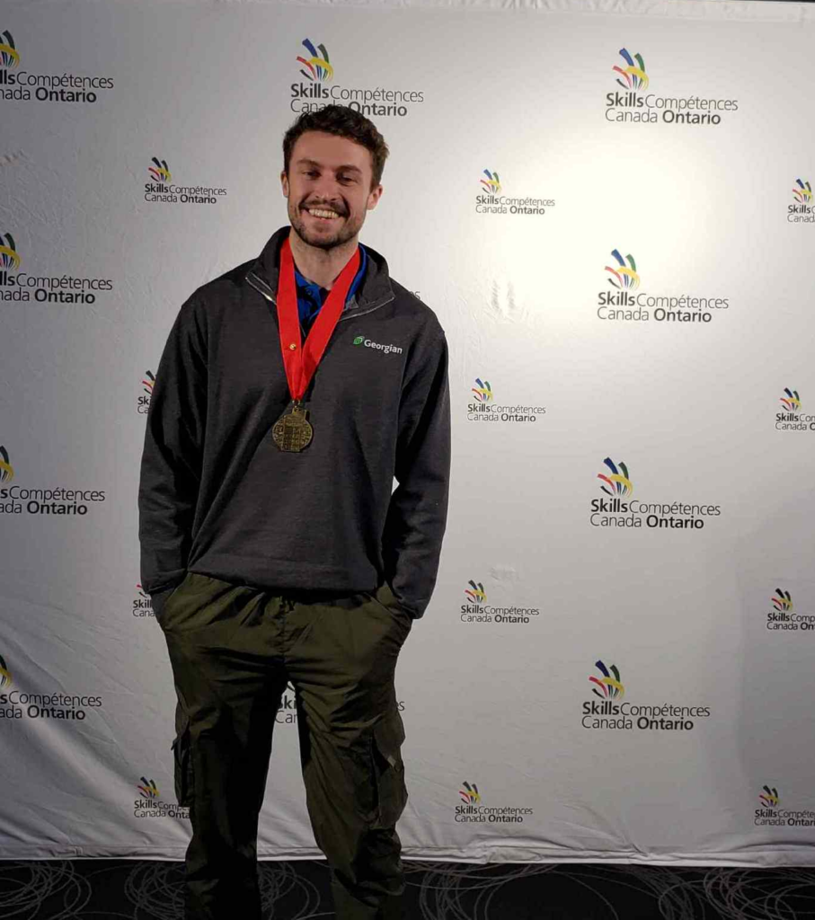 A person wearing a grey sweater and green pants stands in front of a white backdrop with a gold medal.