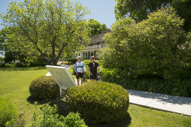 Two bikers on a trail looking at a plaque; trees and open area around them, resort in the background