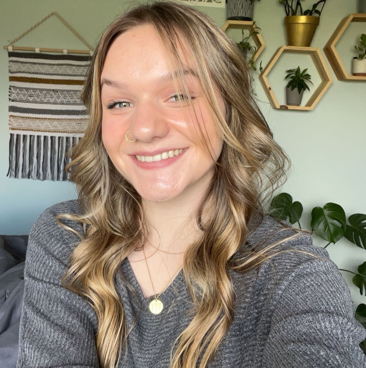 Brooke Park, a student in the inaugural cohort of Georgian's new RPN-to-RN nursing degree bridge smiles broadly at the camera. Her long blond hair is worn down in waves and she's wearing a grey long-sleeved top. She has a nose ring and is wearing minimal makeup. She is sitting in her living room. 