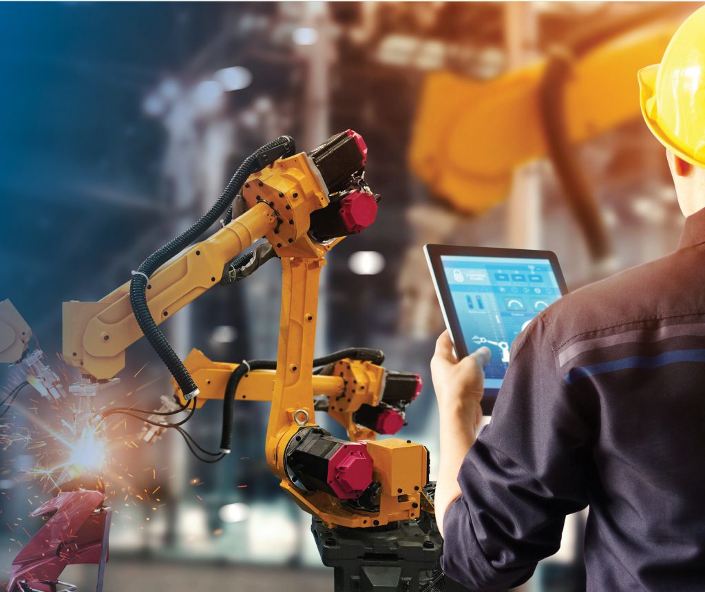 A person in a manufacturing plant, holding a tablet, standing in front of a robotic arm