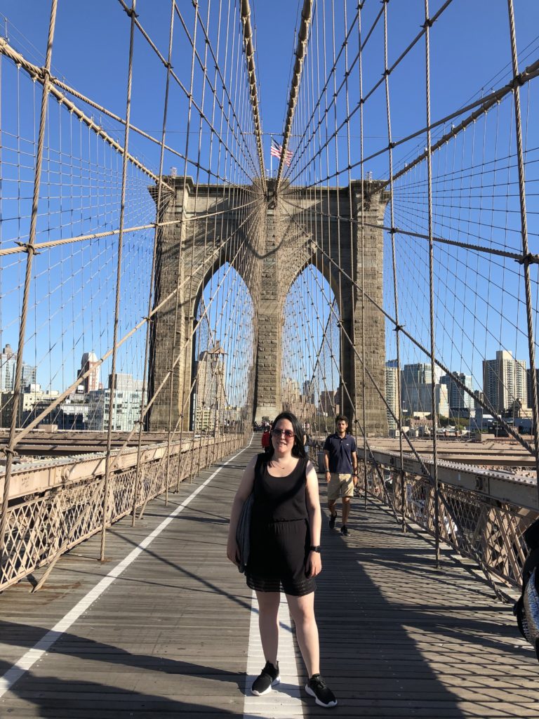 A person stands outside and smiles on a large bridge.
