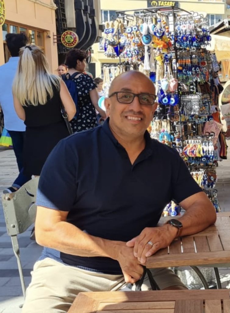 A person with glasses, blue shirt and tan pants, sits at an outdoor table in front of a vendor market.