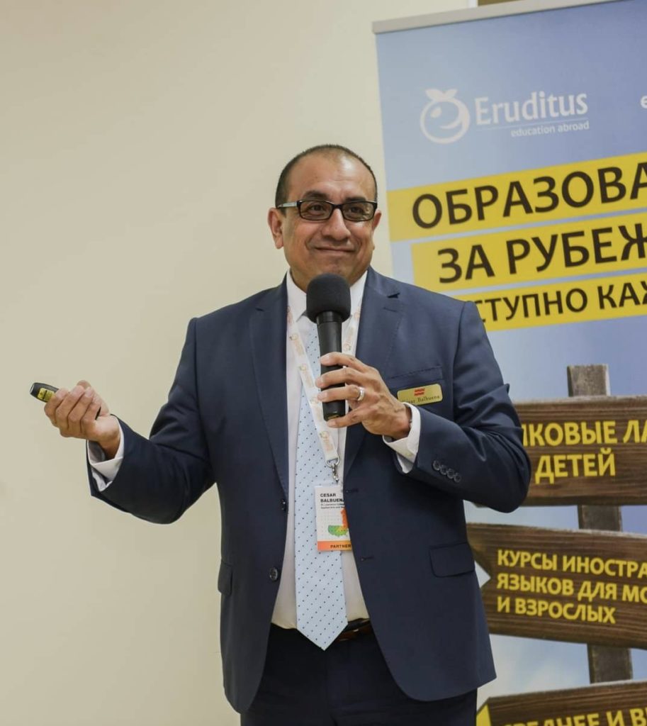 A person with glasses and wearing a suit and tie, smiles and holds a microphone.