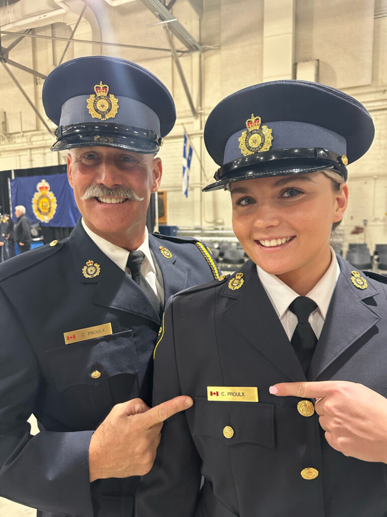 A man and a woman stand together wearing navy blue police uniforms and hats, smiling. 