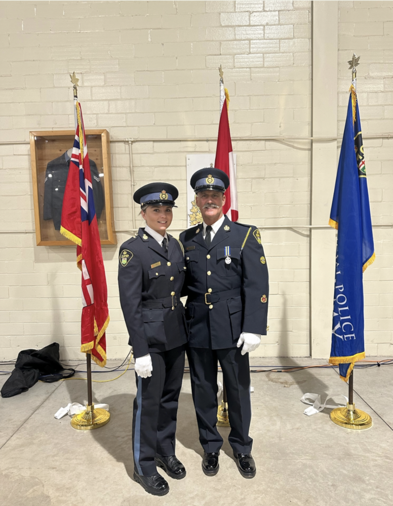 A man and a woman stand together wearing navy blue police uniforms and hats, smiling. Flags are in the background.