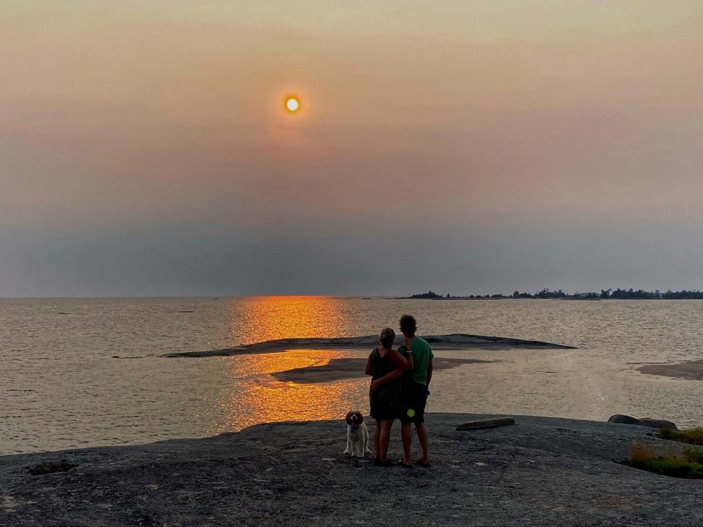 Two people wearing shorts and T-shirts stand with their arms around each other, and a dog sitting at their feet, as they look out at a sunset on a lake.