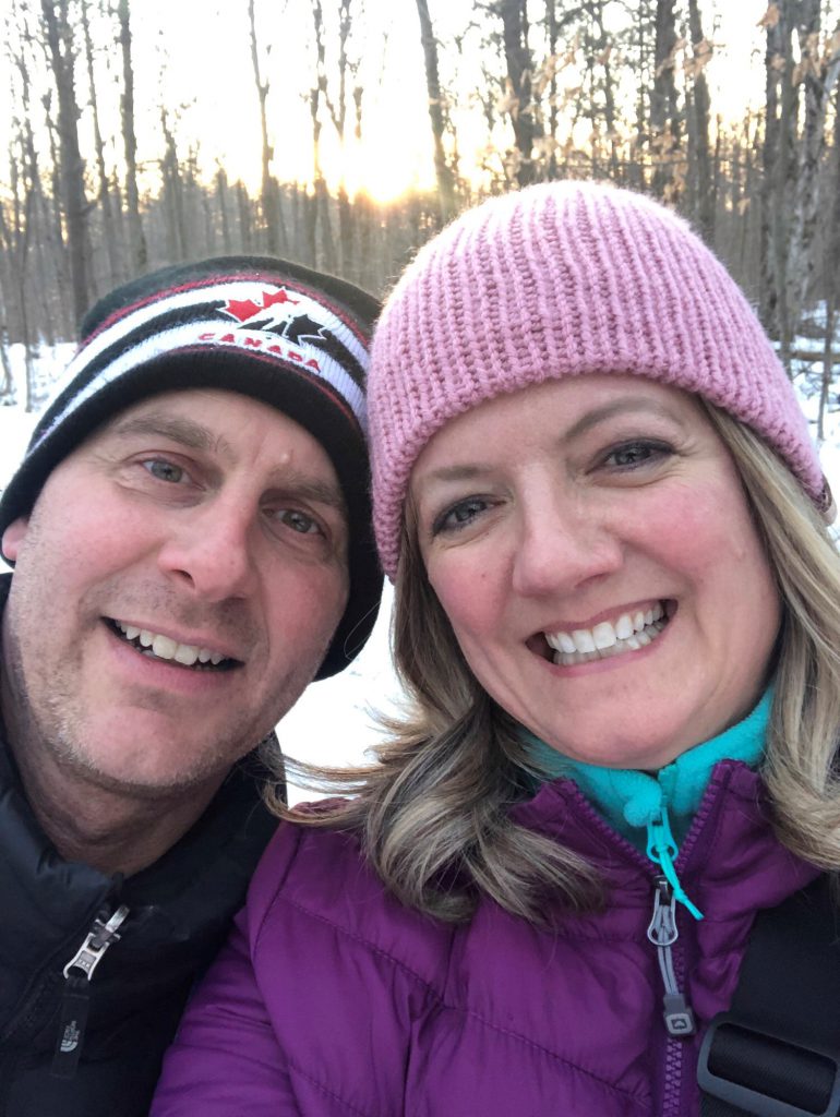 Two people dressed in winter hats and coats stand together and smile at the camera outside in a snowy forest.