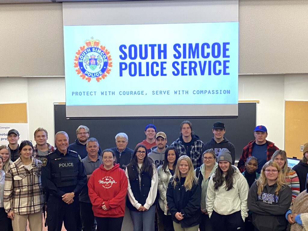 A large group of students from Georgian College community safety programs stand in a classroom with the South Simcoe Police Service logo on a screen behind them. 