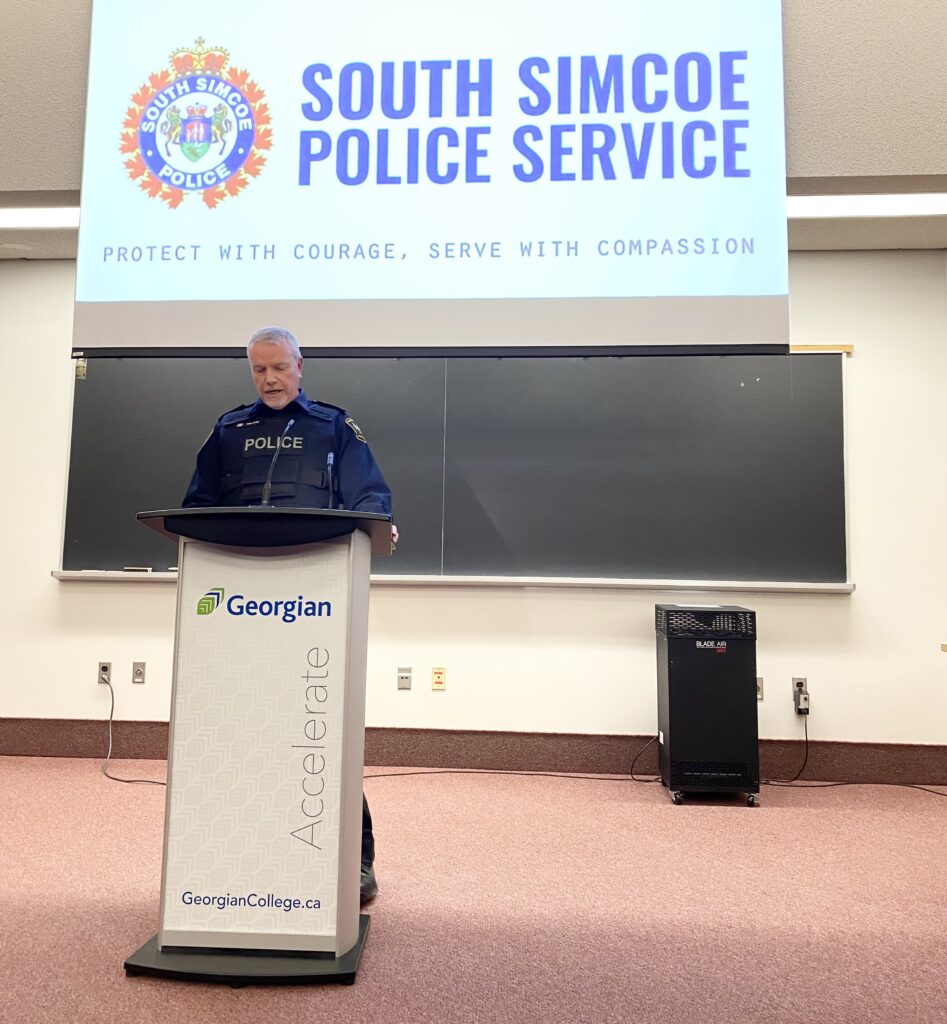 A man in a policing uniform stands at a white Georgian College podium in a classroom with the South Simcoe Police Service logo on a screen behind him.