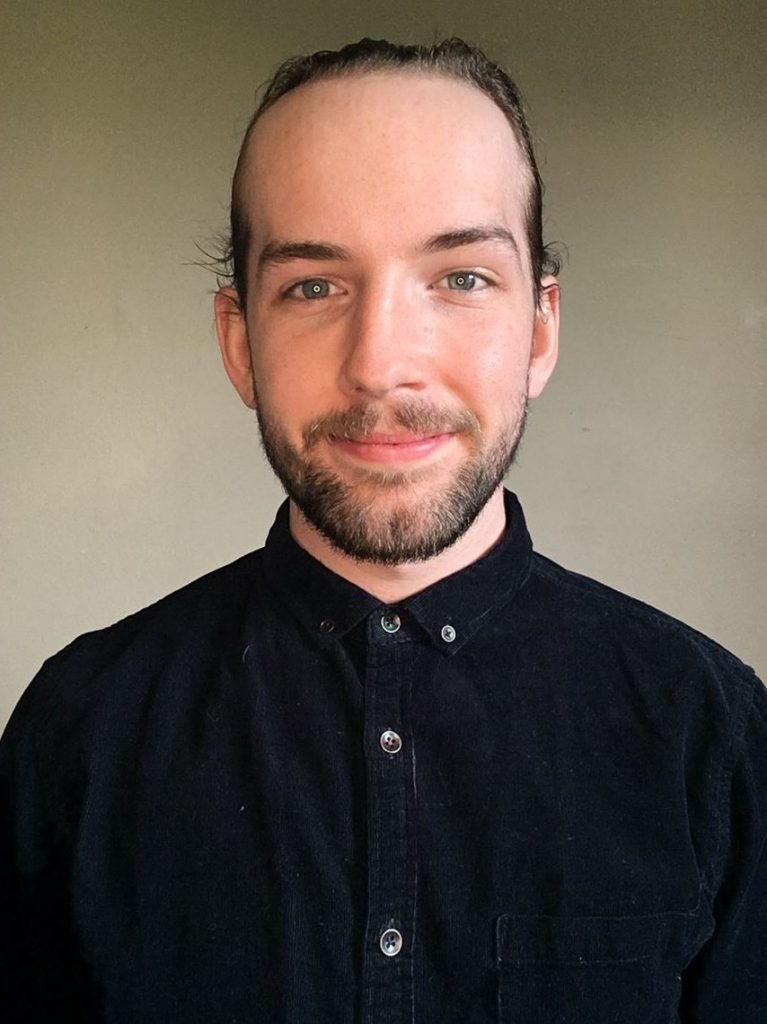 A person with brown hair pulled back and a brown beard, and who's wearing a black buttoned shirt, smiles at the camera.