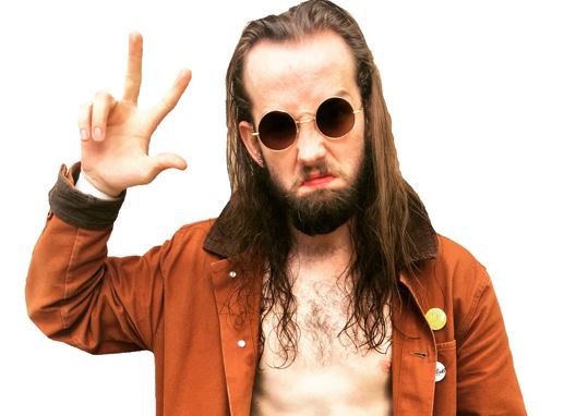 A person with long brown hair and beard, circular sunglasses, shirtless, and orange jacket, grimaces while holding up three fingers on one hand.