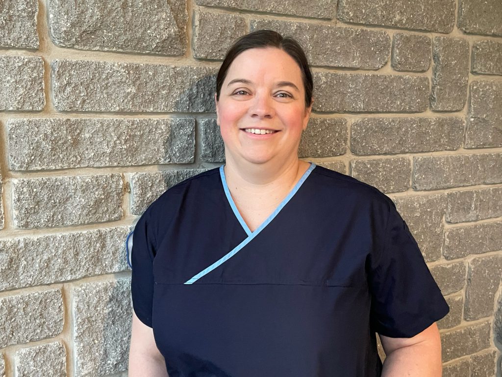 A person wearing scrubs smiles at the camera while standing against a brick wall.