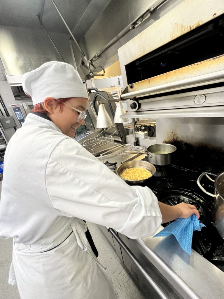 A student wipes down a stove burner, and they're wearing a white chef's hat and coat.