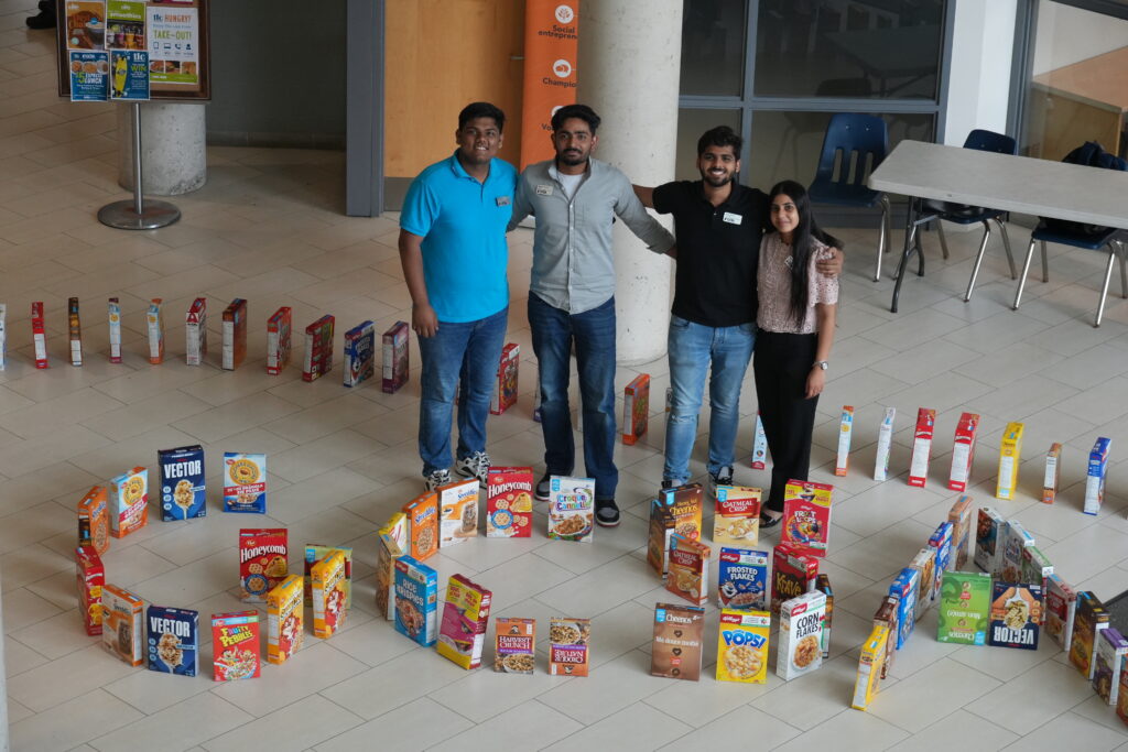 Four people stand together next to boxes of cereal set up in a line and spelling out the letters GCSA.