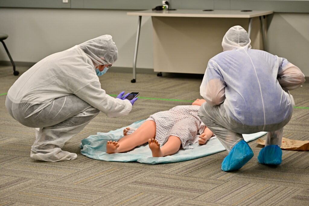 Two students wearing protective clothing examine a mock crime scene, one takes a photo.