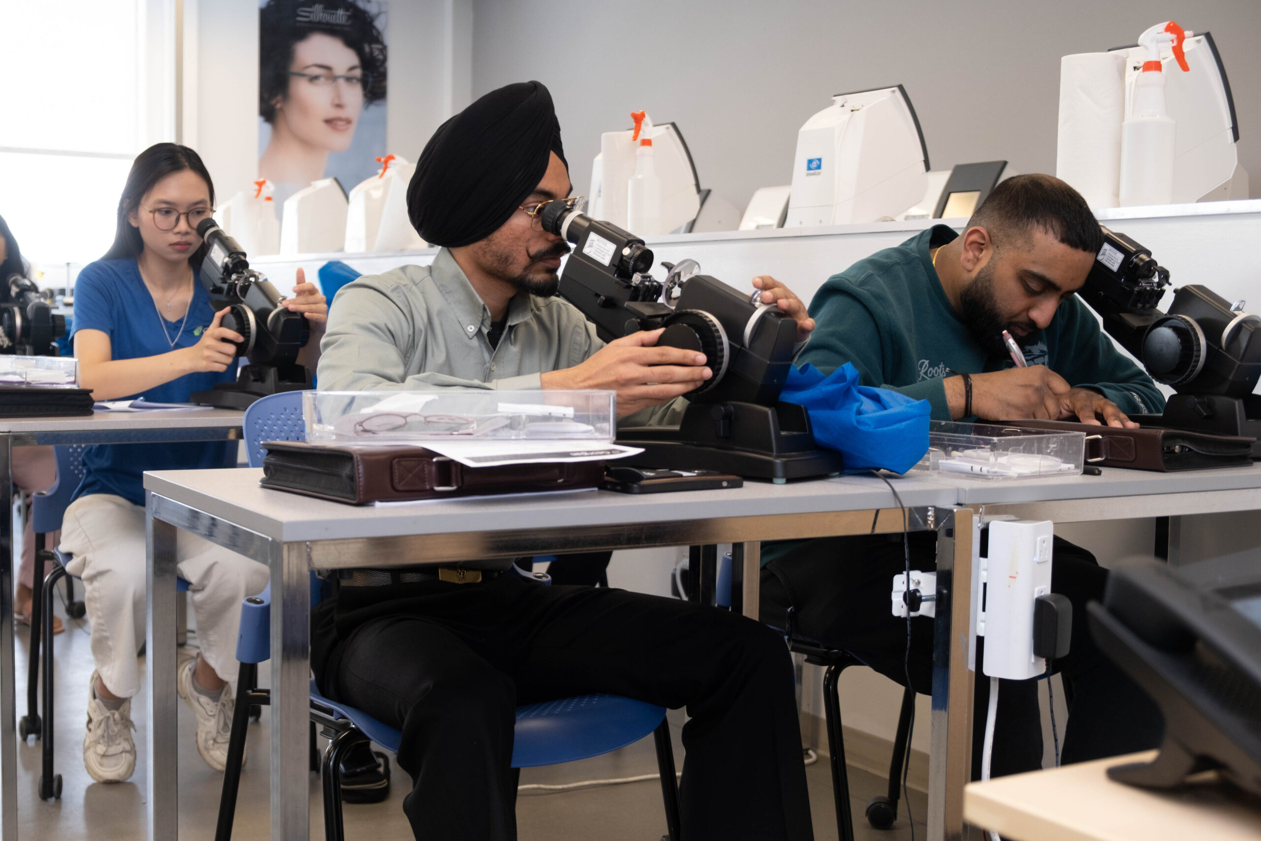 Students in a classroom look through a piece of optometry equipment.