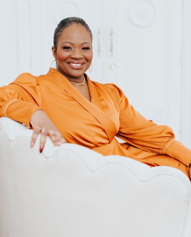 A person with their hair pulled back, wearing gold hoop earrings and an orange dress, smiles and sits on a white chair.