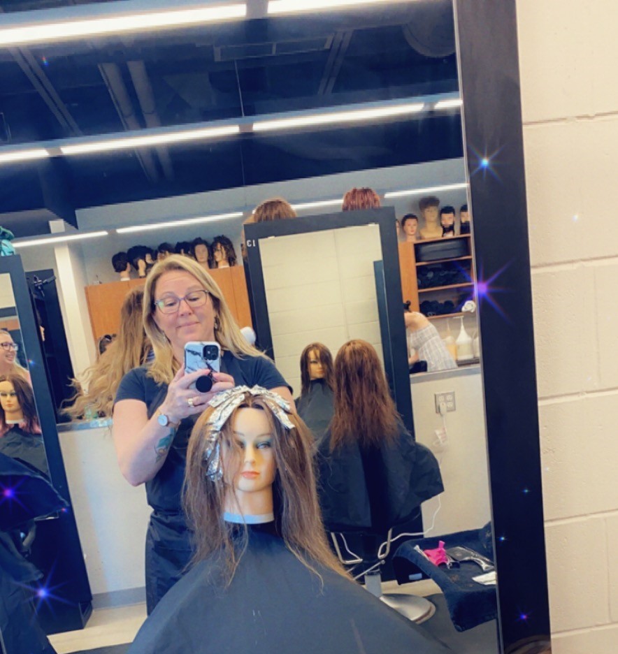 A woman with blonde hair takes a selfie in a mirror with a mannequin head while she styles its hair in a hair salon.