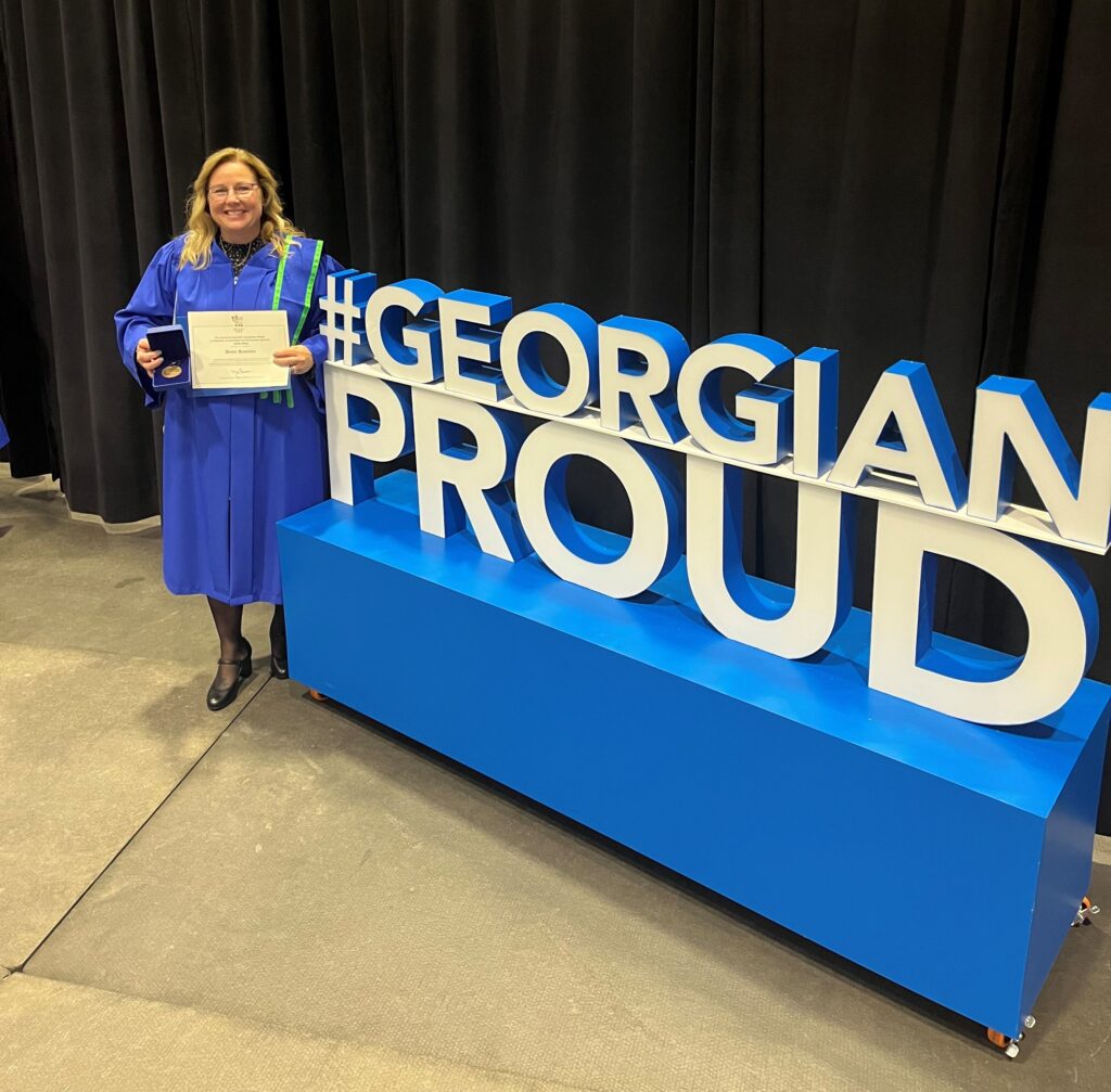 A woman with blonde hair wears a blue graduation gown and stands next to a sign that says "#GeorgianProud" and she holds a certificate and a medal.