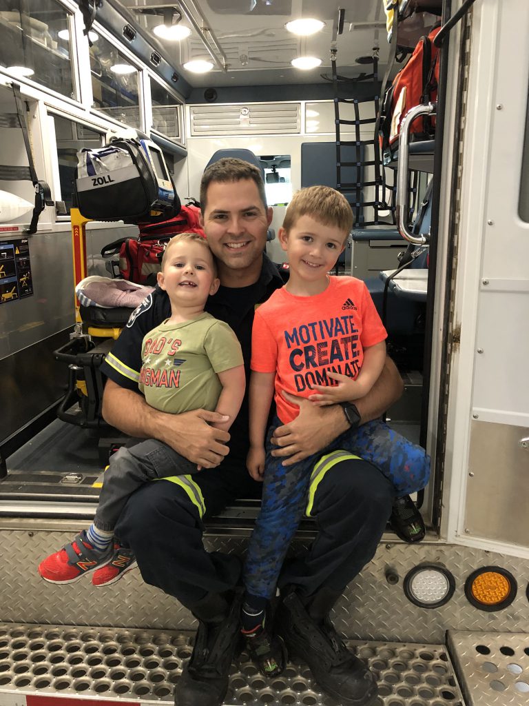 A person with brown hair and dressed in a paramedic uniform sits in the back of an ambulance with two children on their lap, all of them smiling at the camera.