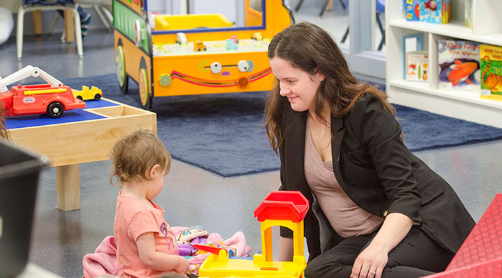 A woman sitting on the floor watching a toddler play with a yellow play garage