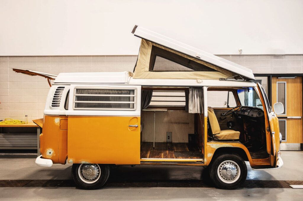 Side view of an orangey yellow Volkswagen Westfalia camper van that is parked inside a workshop with its doors open.