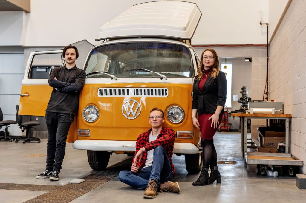Two people stand and one person sits next to a Volkswagen Westfalia camper van that is parked inside a workshop.