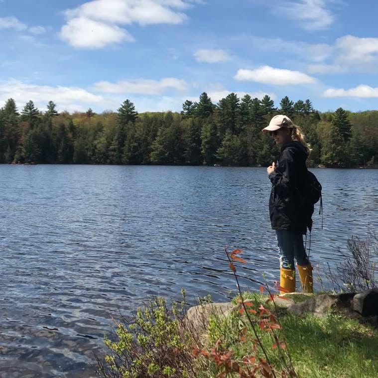 A woman stands at the edge of a lake wearing rubber boots.