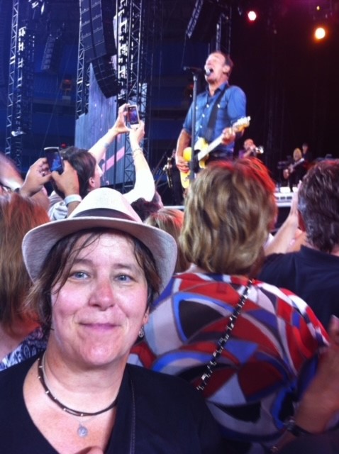 A person takes a selfie in a concert crowd next to a stage where Bruce Springsteen is performing.