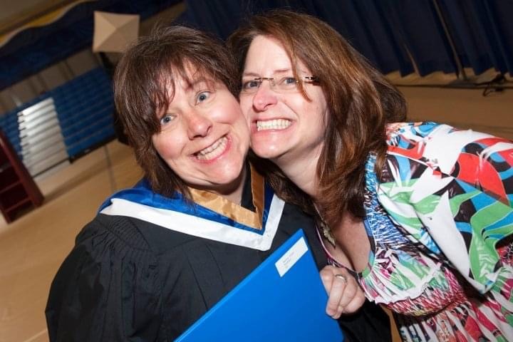 Two people, one of whom is wearing a graduation gown, smile with their heads touching.