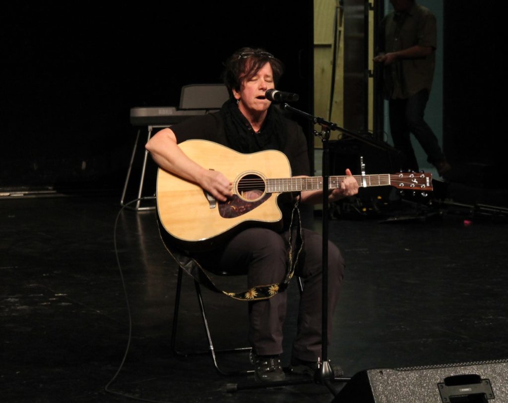 A person dressed in black sits on a chair and sings a song while playing the guitar.
